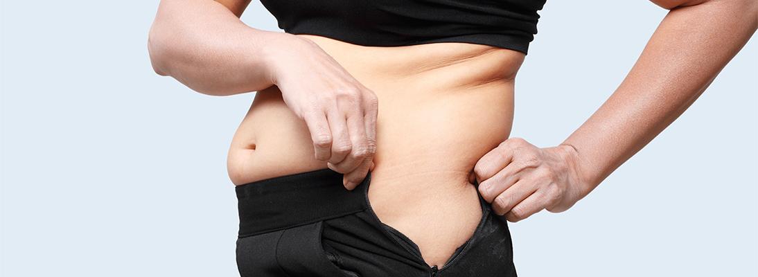 Femme montrant de la graisse abdominale et essayant de fermer un pantalon serré.